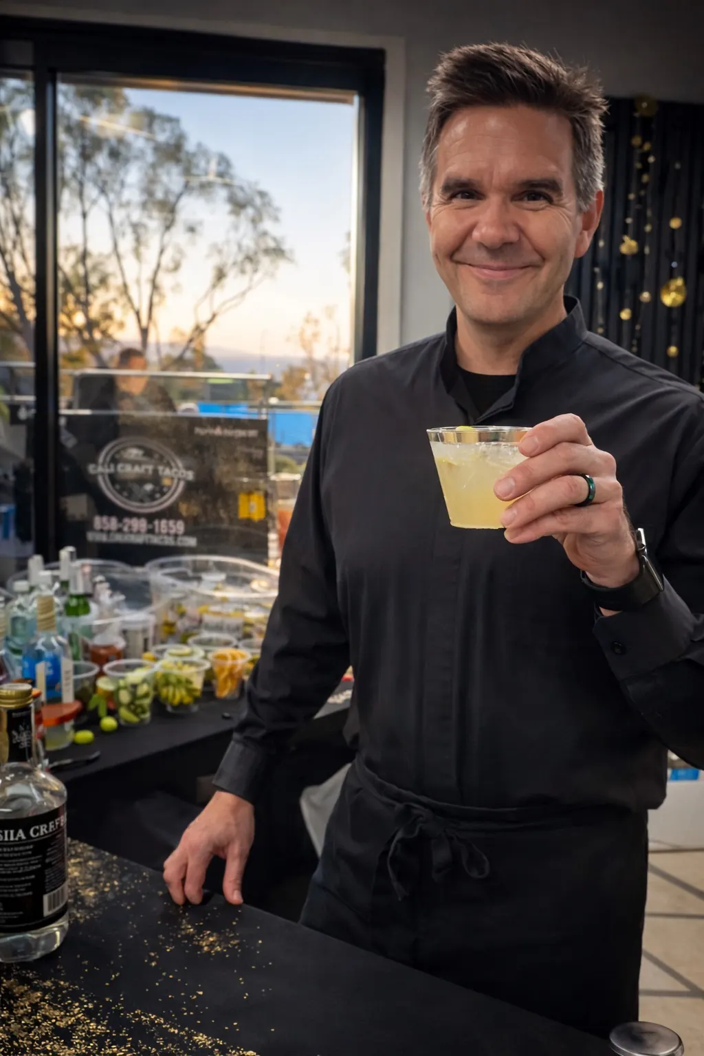 Justin in a black bartender shirt raising a cocktail at an evening event bar
