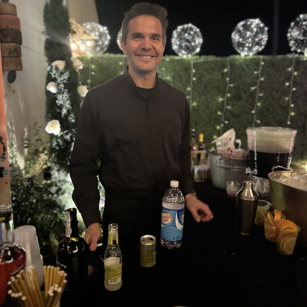 Justin behind a night event bar with glowing light globes and a green hedge wall
