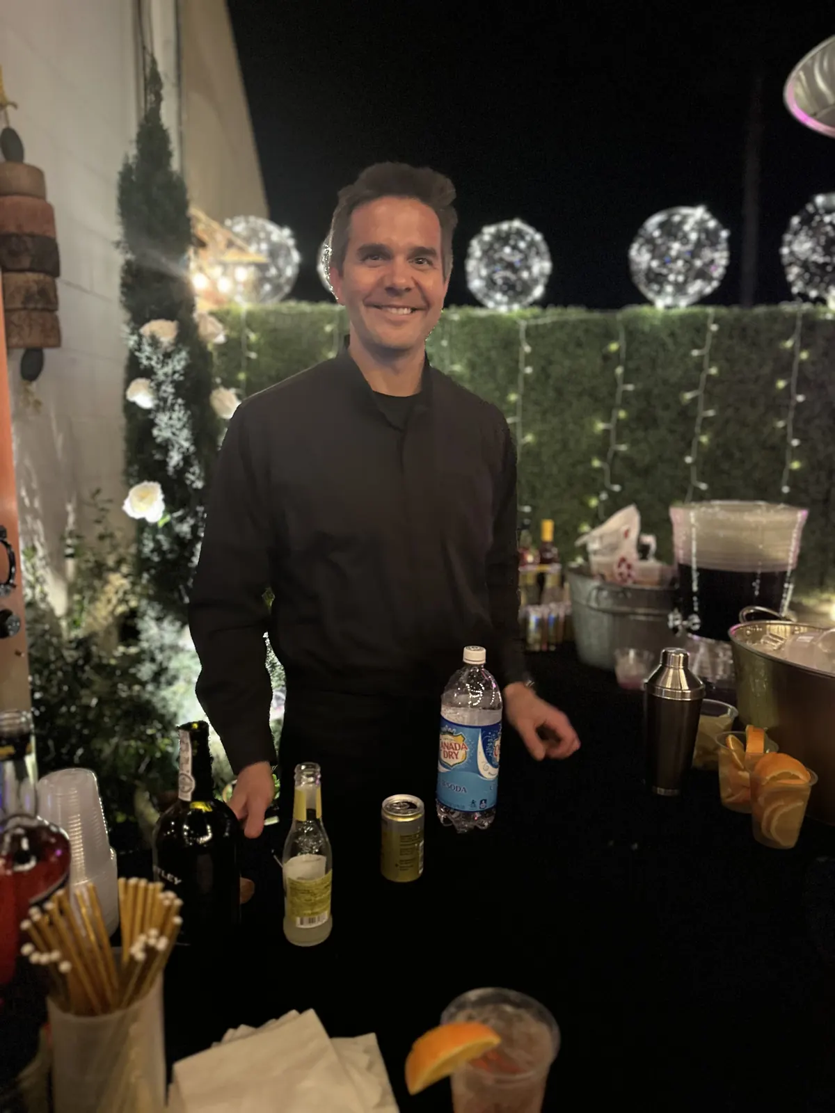 Justin behind a night event bar with glowing light globes and a green hedge wall