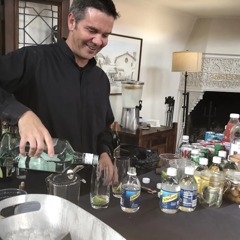 Justin pouring a clear spirit into a glass with lime at an indoor event bar lined with mixers
