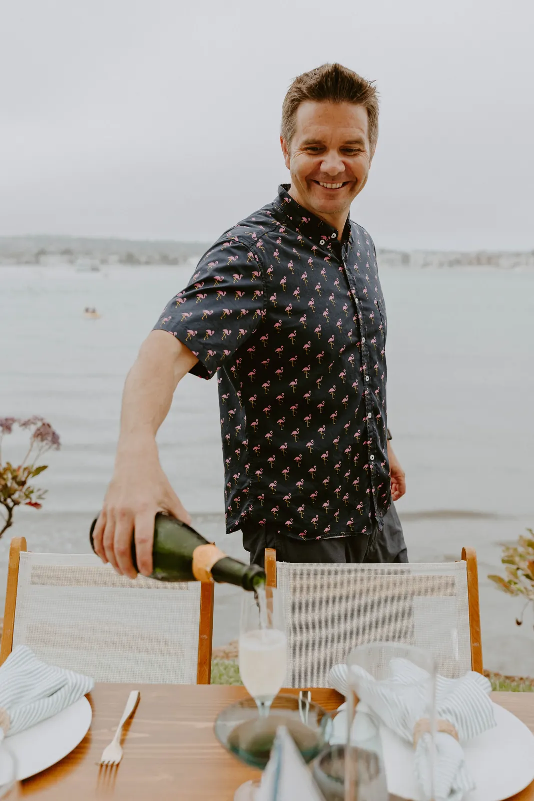 Justin pouring sparkling wine into a glass at a set dinner table beside the water