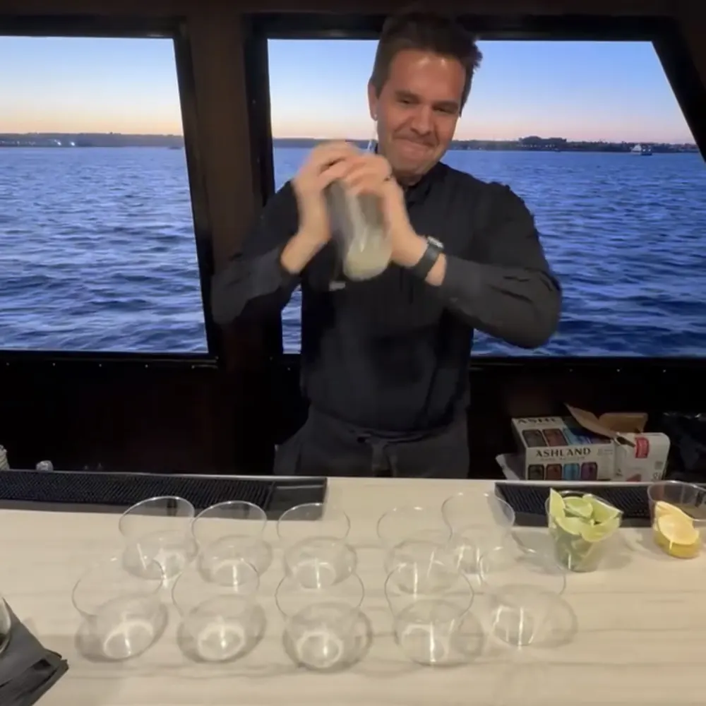 Justin shaking a cocktail behind a boat bar with the water and a sunset sky in the windows
