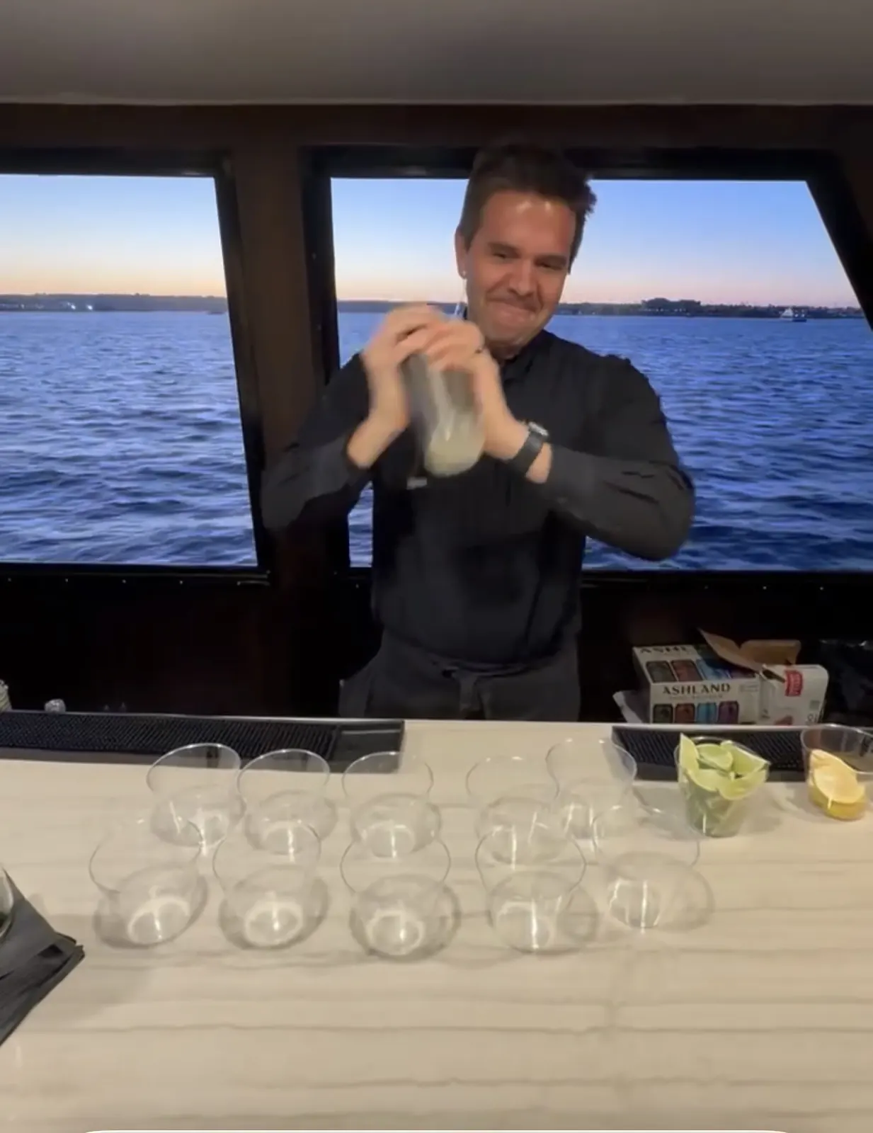 Justin shaking a cocktail behind a boat bar with the water and a sunset sky in the windows