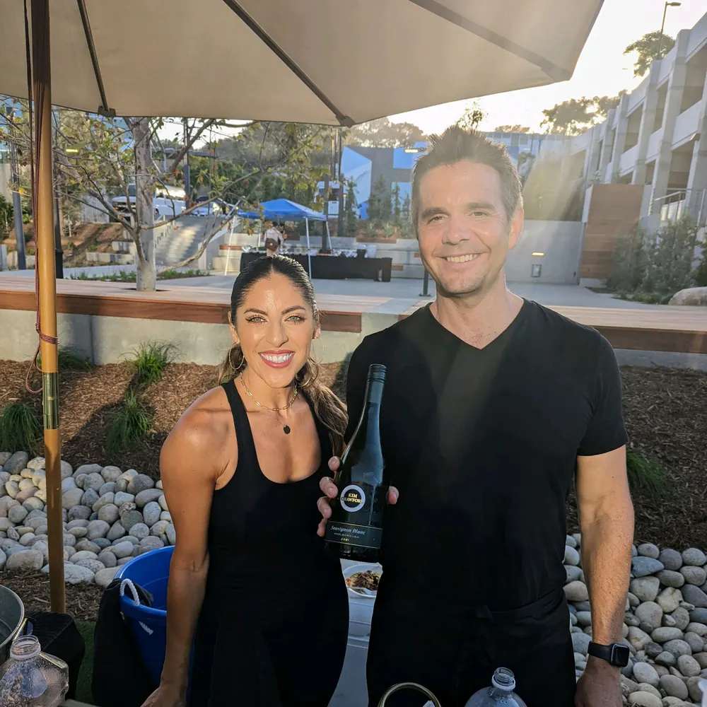Justin holding a bottle of white wine next to a smiling woman at a sunny outdoor event bar