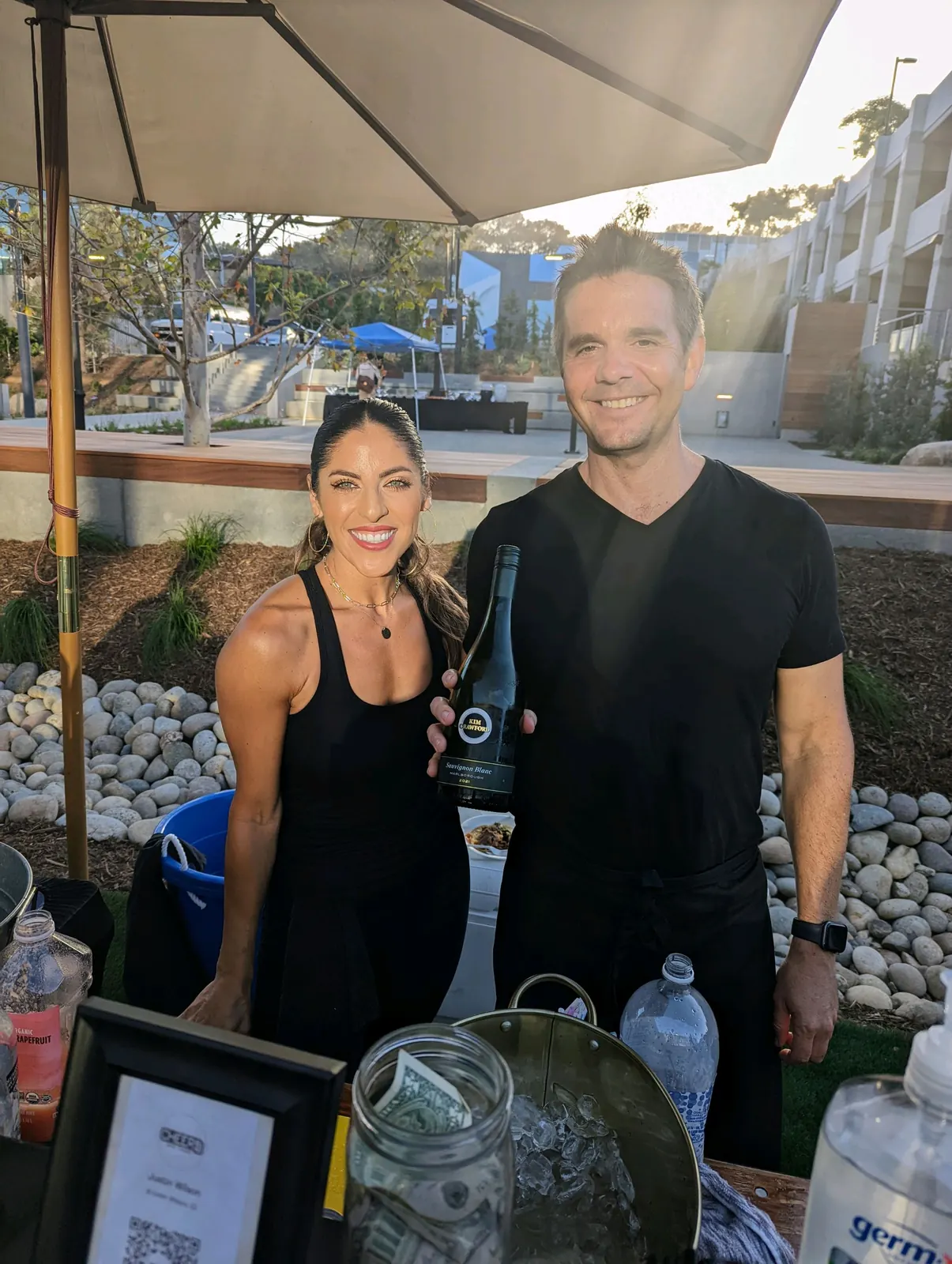 Justin holding a bottle of white wine next to a smiling woman at a sunny outdoor event bar