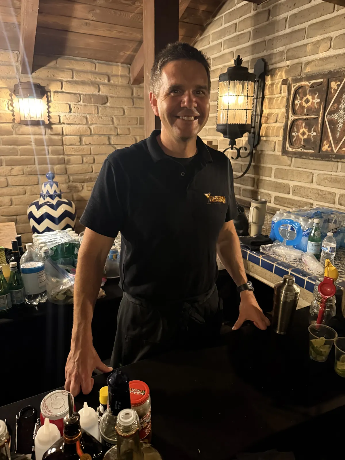 Justin smiling behind the bar at a night event, brick wall and lanterns behind him