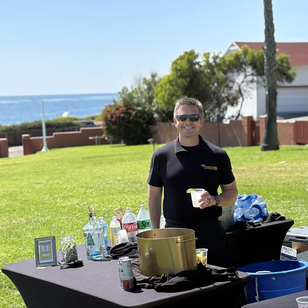 Justin in a Cheers San Diego polo holding a drink behind a mobile bar on a lawn with the ocean behind