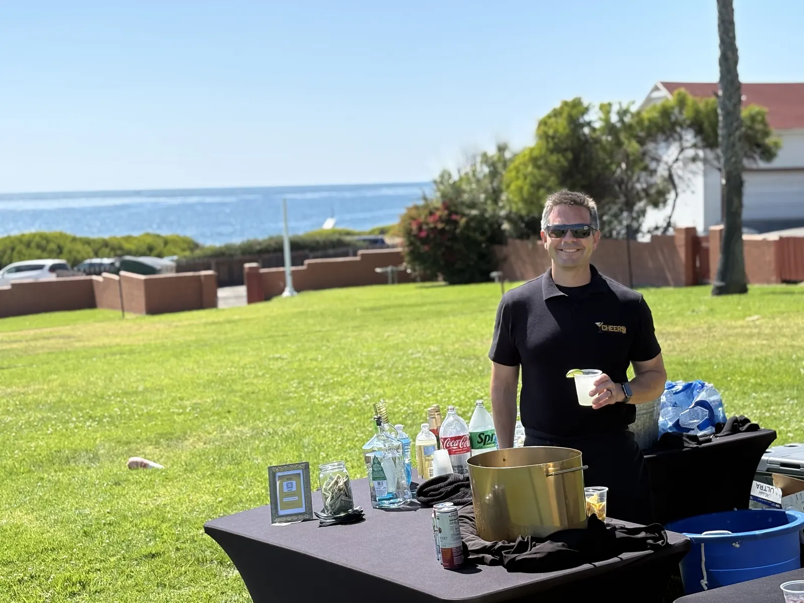 Justin in a Cheers San Diego polo holding a drink behind a mobile bar on a lawn with the ocean behind