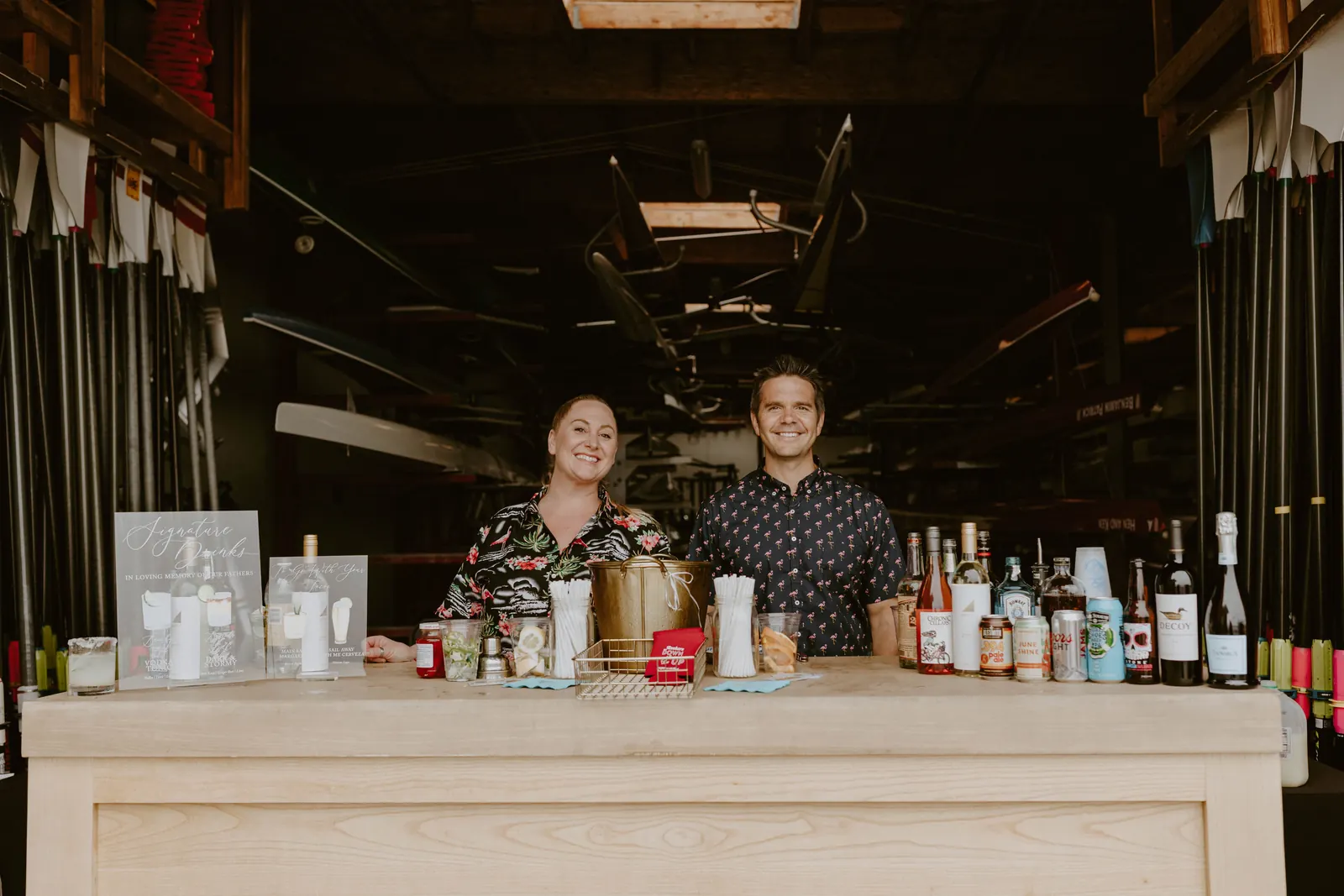 Two smiling bartenders behind a light wood mobile bar set with signature drink signs, bottles and an ice bucket